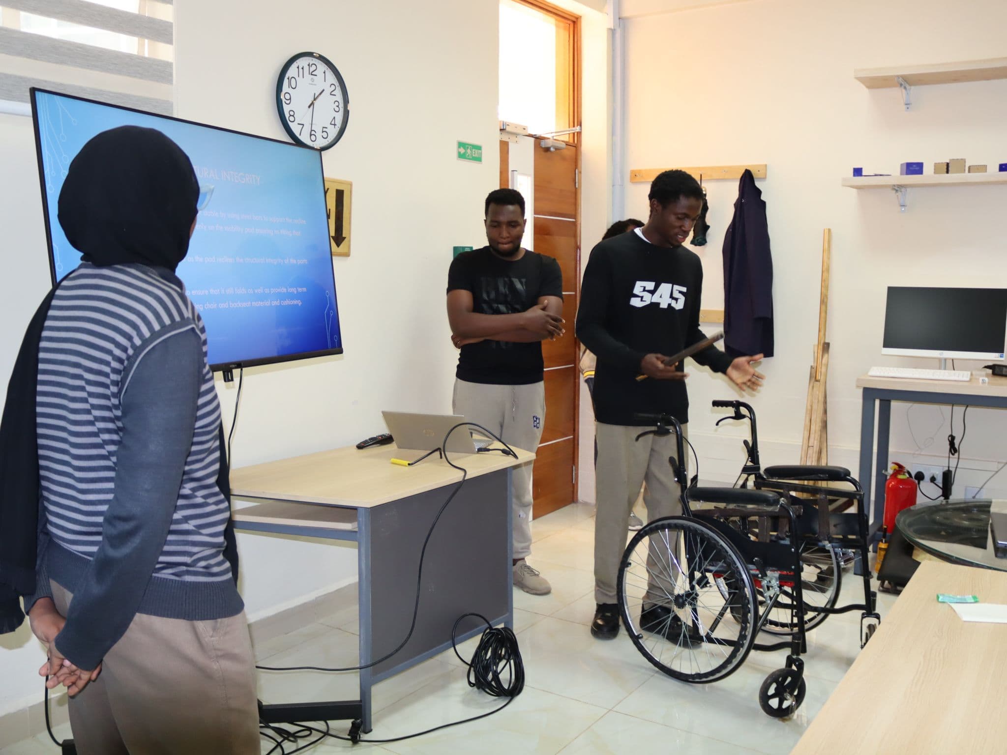 A team pitching a reclining wheelchair concept beside two wheelchairs, with a slide on structural integrity