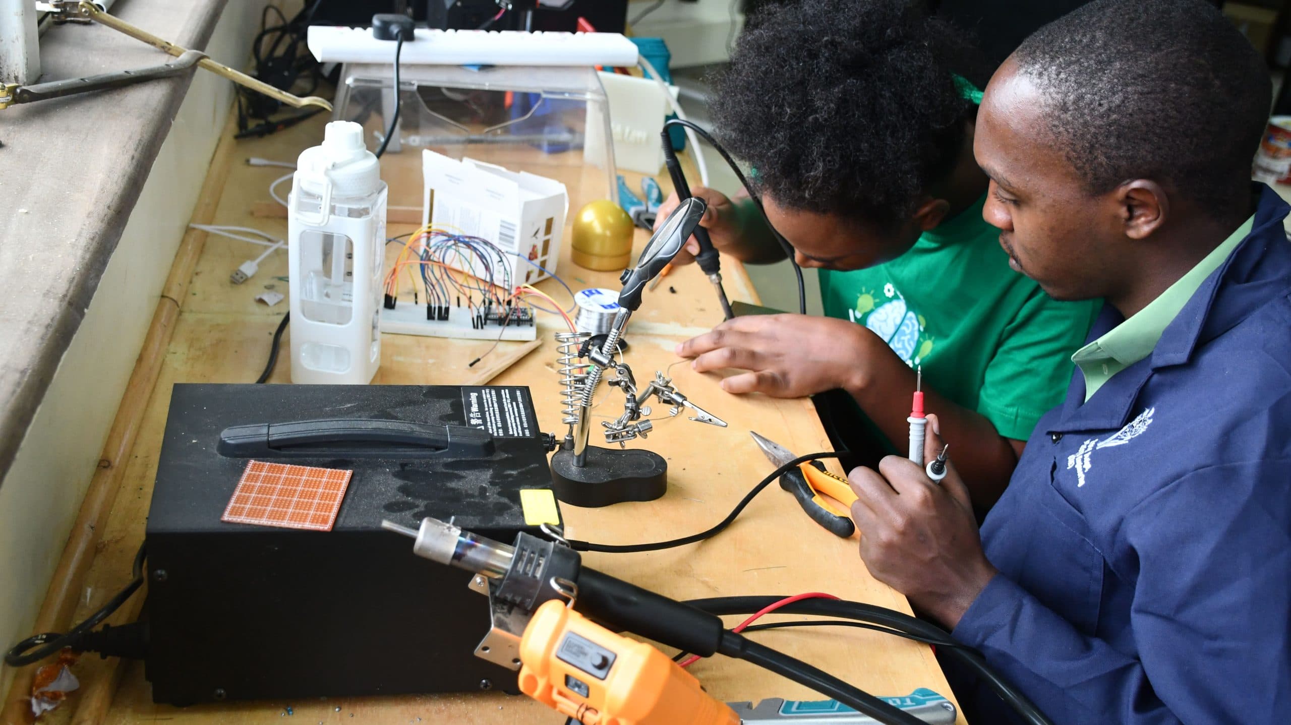 Two students at a bench soldering with a helping-hands clamp beside a breadboard
