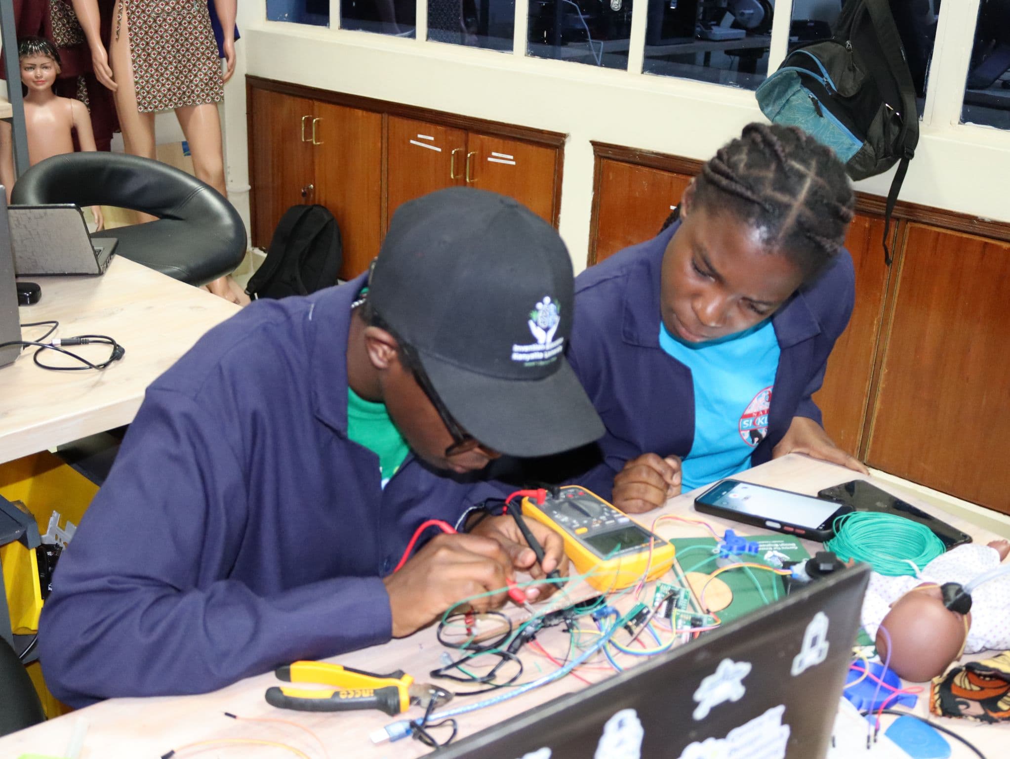 Two participants testing a circuit with a multimeter beside an infant training mannequin